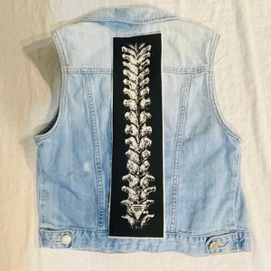 Op de afbeelding: Een lichtblauw denim vest met een zwart-witte wervelkolom grafische patch op de rug.