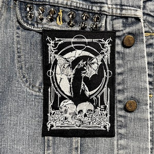 May include: Black and white embroidered patch with a bat, skulls, and a pentagram design. The patch is on a denim jacket.