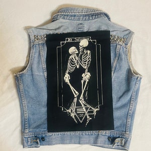 May include: Light blue denim vest with a distressed hem and silver stud embellishments on the shoulders. A large black patch on the back features a white illustration of two skeletons embracing, with the text "AD INFINITUM" above and a geometric design below.