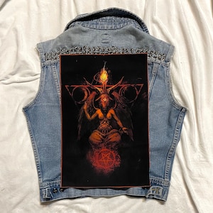 Peut inclure: Veste en jean bleu clair avec un grand écusson rectangulaire représentant Baphomet. La veste est ornée de clous argentés le long du col. L'image de Baphomet est dans les tons de rouge, orange et noir.