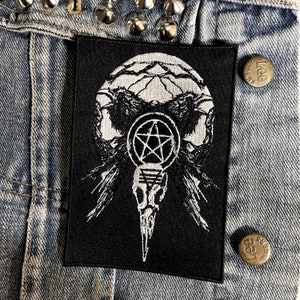 Op de afbeelding: Zwart-witte geborduurde patch met twee raven, een pentagram en een schedel met een lange snavel. De patch is op een spijkerjack.