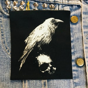 May include: A black iron-on patch featuring a white silhouette of a raven perched on a skull.