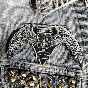 May include: A black and white embroidered patch of a gas mask with wings. The patch is sewn onto a denim jacket.