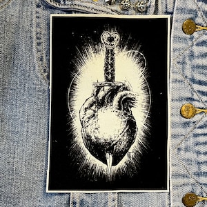 Puede incluir: Parche bordado en blanco y negro de un corazón con una daga que lo atraviesa. El corazón está rodeado por un halo blanco.