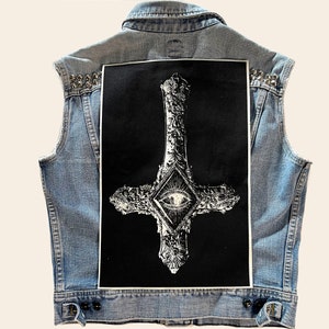 May include: A light blue denim vest featuring silver metal studs on the shoulders and a large rectangular black patch on the back. The patch displays a white, ornate, inverted cross design with a central diamond containing an eye graphic.