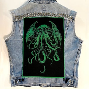May include: A light blue denim vest with silver studs along the shoulders. The back features a large black patch with a green illustration of a creature with tentacles and wings.