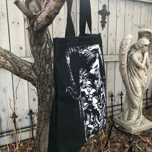 May include: Black tote bag with a white graphic print of a creature with horns and wings. The bag has black straps and a white lining.
