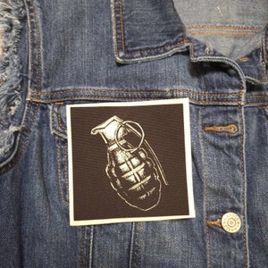 May include: A black and white embroidered patch of a grenade with a pull pin on a denim jacket.
