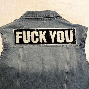 Könnte beinhalten: Eine ärmellose Jeansjacke mit einem großen schwarz-weißen Aufnäher auf dem Rücken. Der Aufnäher zeigt die Worte "FUCK YOU" in weißen Großbuchstaben.