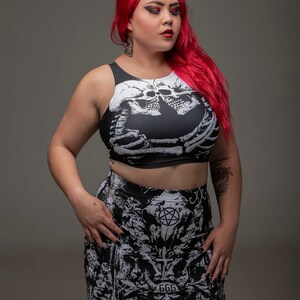 May include: A woman with long red hair is wearing a black and white crop top with a skull and skeleton design. She is also wearing a black and white mini skirt with a pentagram and other gothic imagery. The skirt has the text "666" and "Mayhem" printed on it.