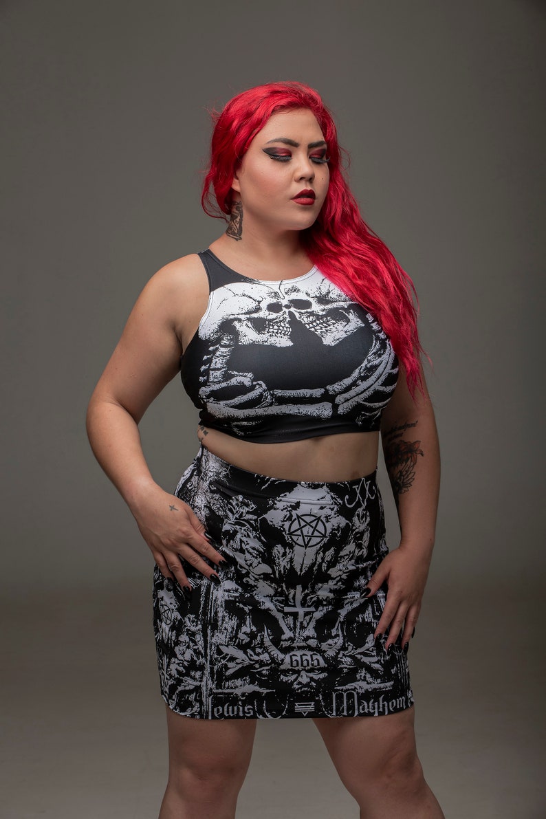May include: A woman with long red hair is wearing a black and white crop top with a skull and skeleton design. She is also wearing a black and white mini skirt with a pentagram and other gothic imagery. The skirt has the text "666" and "Mayhem" printed on it.