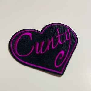 May include: A black heart-shaped patch with the word "Cunty" embroidered in bright pink. The patch has a pink border and is likely made of fabric.