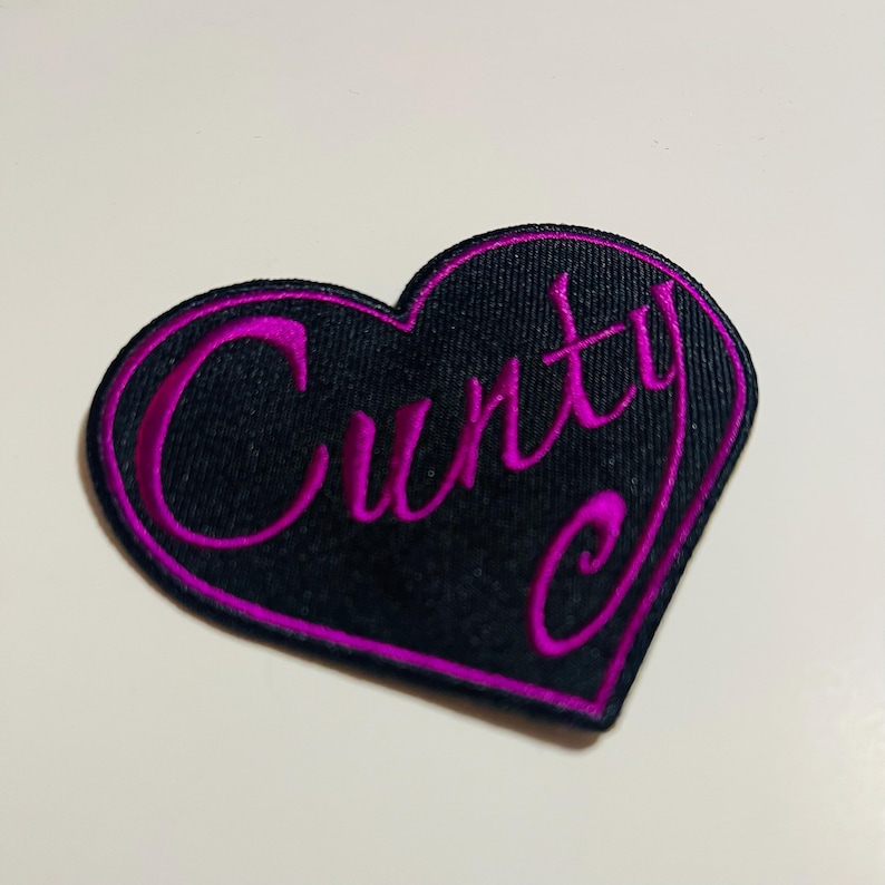 May include: A black heart-shaped patch with the word "Cunty" embroidered in bright pink. The patch has a pink border and is likely made of fabric.