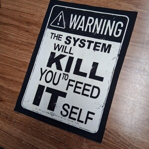 May include: A black and white rectangular patch with the text "WARNING THE SYSTEM WILL KILL YOU TO FEED IT SELF". The patch has a black border and a warning symbol at the top. The text is in a bold, distressed font.