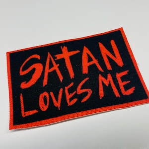 May include: Red embroidered patch with a black background. The patch reads "SATAN LOVES ME" in a bold, handwritten style.