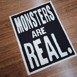 May include: A black and white rectangular sign with the words "MONSTERS ARE REAL" printed in a bold, sans-serif font. The text is white against a black background. The sign is placed on a wooden surface.
