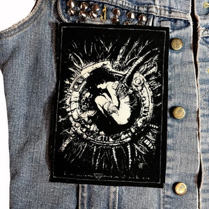 Può includere: Toppa ricamata in bianco e nero con un design dettagliato di una figura umana intrappolata all'interno di una struttura meccanica circolare. La toppa ha un aspetto usurato e vintage, perfetta per aggiungere un tocco unico a una giacca di jeans o ad altri indumenti.