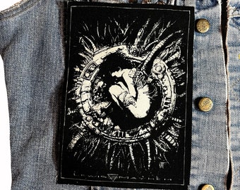 May include: Black and white embroidered patch featuring a detailed design of a human figure trapped within a circular, mechanical structure. The patch has a distressed, vintage aesthetic and is ideal for adding a unique touch to a denim jacket or other clothing.