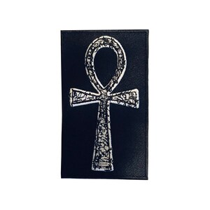 Puede incluir: Un parche de tela negro con un s&iacute;mbolo Ankh bordado en blanco.