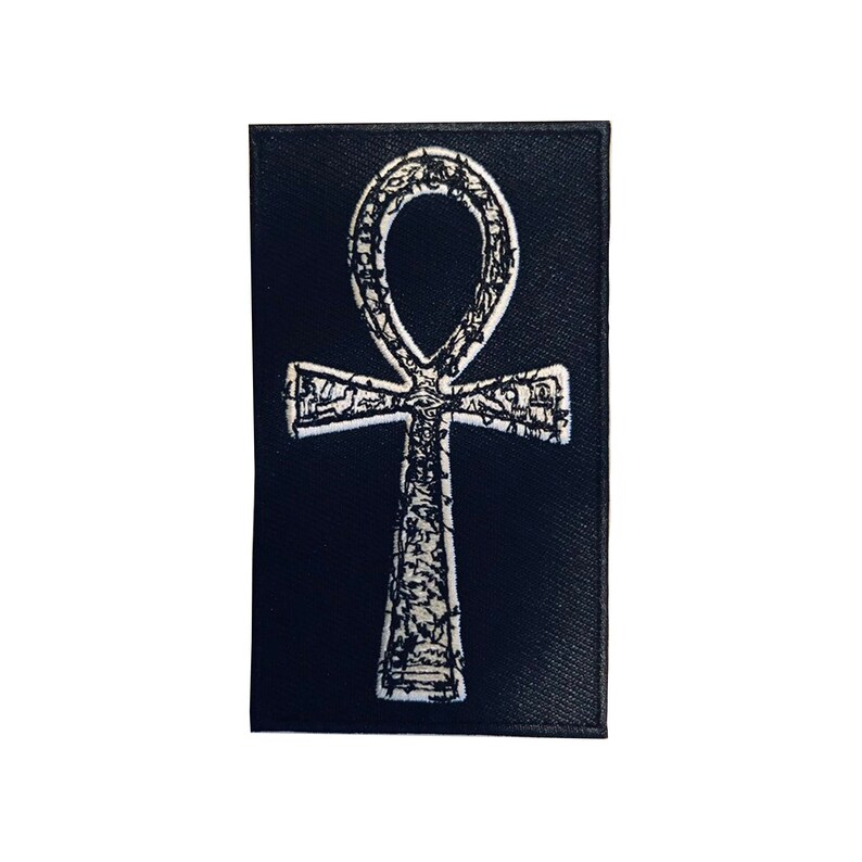 Puede incluir: Un parche de tela negro con un s&iacute;mbolo Ankh bordado en blanco.