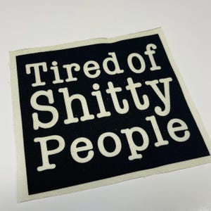 May include: A square black and white patch with the text "Tired of Shitty People" in bold, white capital letters. The patch has a white border and is likely made of fabric.