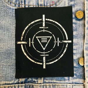 Può includere: Toppa ricamata in bianco e nero con un simbolo di bersaglio e un triangolo con tre linee all'interno. La toppa ha un aspetto usurato.