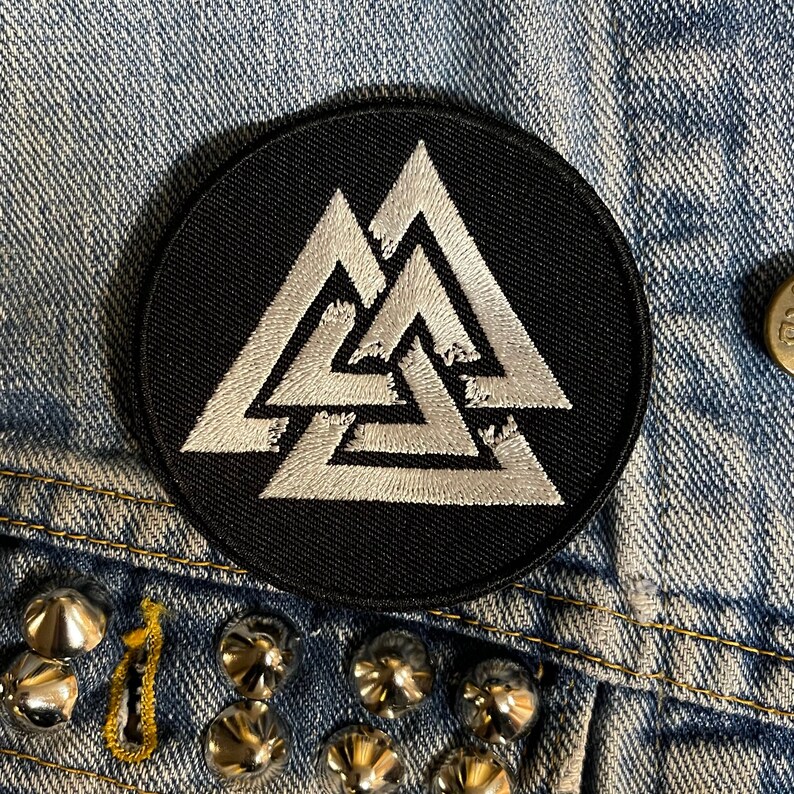 May include: A black and white embroidered patch featuring a triple triangle symbol, also known as the Valknut, on a black circular background.