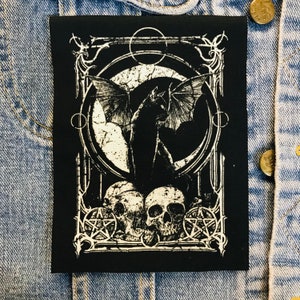 May include: Black and white embroidered patch featuring a bat-like creature with wings outstretched, encircled by a circle with a crescent moon and stars. The creature stands on two skulls with pentagrams on either side.