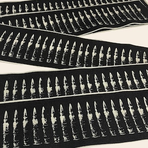 Könnte beinhalten: Schwarzer Stoff mit einem sich wiederholenden Muster aus weißen Patronenhülsen. Der Stoff ist dreifach gefaltet und zeigt drei Reihen des Musters.