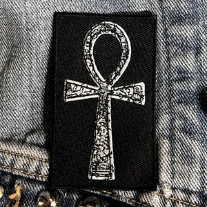 May include: A black patch with a white Ankh symbol. The symbol is detailed with a textured pattern.