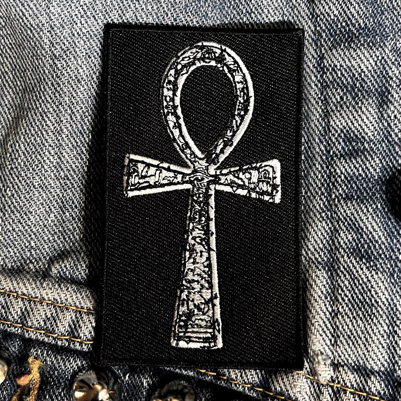 Puede incluir: Un parche negro con un s&iacute;mbolo Ankh blanco. El s&iacute;mbolo est&aacute; detallado con un patr&oacute;n texturizado.