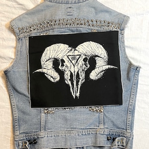 Op de afbeelding: Een denim vest met zilveren studs en een zwarte patch met een witte schedel met hoorns en een driehoek symbool.