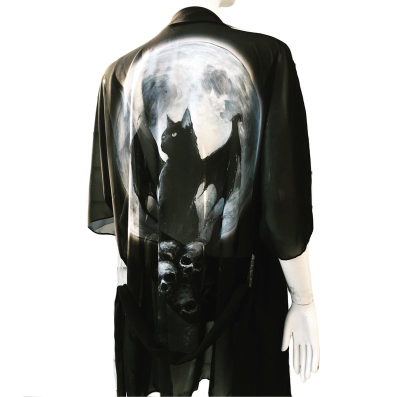 May include: Black chiffon kimono with a moon and a black cat with wings design. The cat is perched on a skull with other skulls below it.
