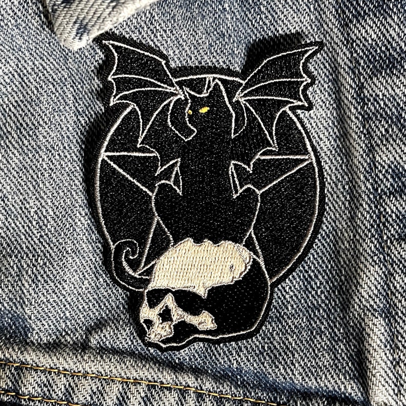 Pode incluir: Etiqueta bordada preta de um gato com asas de morcego sentado em um cr&acirc;nio com um pentagrama no fundo.