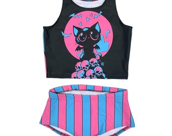 Op de afbeelding: Een zwart tanktop met een roze en blauwe maan, vleermuizen en een zwarte kat met blauwe ogen. Het tanktop wordt gecombineerd met roze en blauwe gestreepte shorts.