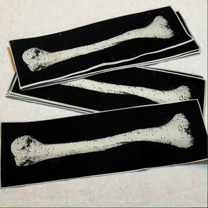 May include: A stack of black fabric patches with a white bone printed on each patch. The bone is a long bone, possibly a femur or humerus.
