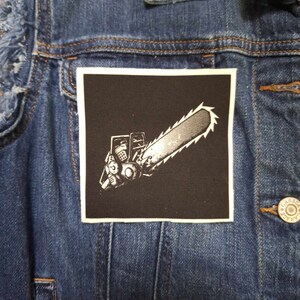 May include: A denim jacket with a black square patch featuring a white chainsaw design. The jacket is a medium wash blue with visible stitching and metal buttons. The patch is centered on the jacket.