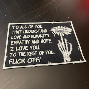 May include: Black rectangular patch with white text and a skeleton hand giving the middle finger. The text reads: "TO ALL OF YOU THAT UNDERSTAND LOVE AND HUMANITY. EMPATHY AND HOPE. I LOVE YOU. TO THE REST OF YOU. FUCK OFF!"