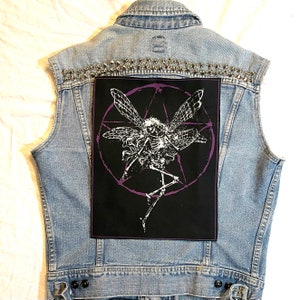 May include: A light blue denim vest with silver stud detailing along the shoulders. A large black patch on the back features a white skeleton fairy design within a purple pentagram.