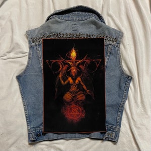 K&ouml;nnte beinhalten: Eine hellblaue Jeansweste mit einem gro&szlig;en Aufdruck auf der R&uuml;ckseite, der eine geh&ouml;rnte Figur mit einer Flamme &uuml;ber dem Kopf vor schwarzem Hintergrund zeigt. Die Weste ist mit silbernen Nieten am Kragen verziert.