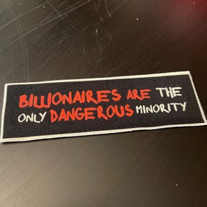 Op de afbeelding: Een rechthoekige zwarte patch met witte rand. De patch bevat de tekst "BILLIONAIRES ARE THE ONLY DANGEROUS MINORITY" in rode en witte letters.