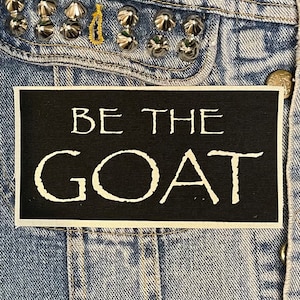 May include: Black and white patch with the text "BE THE GOAT" on a denim jacket.