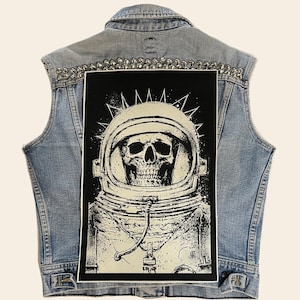 May include: A black denim vest with silver studs and a black and white patch of a skull in a space helmet. The patch has a white background and black details.