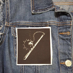 Pode incluir: Um remendo preto e branco com uma imagem branca de um porrete com pontas e uma corrente sobre um fundo castanho.