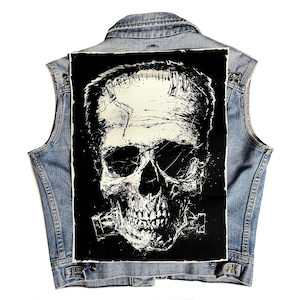 May include: A denim vest featuring a striking black and white skull design on the back. The skull graphic has a distressed, textured appearance. The vest is a light blue wash with a button-up front and classic denim styling.