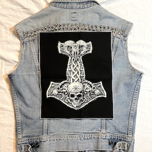 May include: A black and white embroidered patch of a Mjolnir hammer with a skull in the center, surrounded by intricate designs. The patch is sewn onto a denim vest with silver studs.
