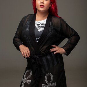 May include: Black sheer robe with an Ankh symbol design and a skull print on the inner lining. The robe has a tie closure at the waist.