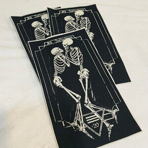 K&ouml;nnte beinhalten: Drei schwarze Stoffpatches mit wei&szlig;en gestickten Skeletten, die sich die H&auml;nde halten. Der Text "Ad Infinitum" befindet sich &uuml;ber den Skeletten. Der untere Patch hat ein Pentagramm mit der Zahl "23" darin.