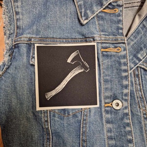 Peut inclure: Veste en jean sans manches avec un écusson d'axe blanc au dos. L'écusson est une illustration blanche d'une hache sur fond noir. La veste a une fermeture boutonnée et un col.