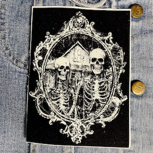 May include: Black and white embroidered patch featuring two skeletons in an ornate frame. The skeletons are standing in front of a house with a window. The frame is decorated with swirls and vines.